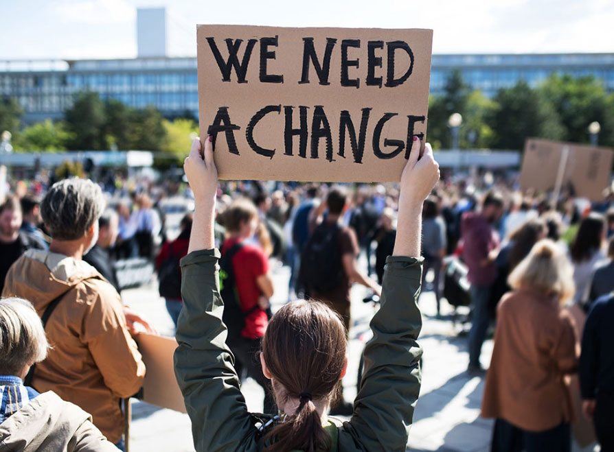 Image of a lady holding up a sign in the street that reads "WE NEED A CHANGE"